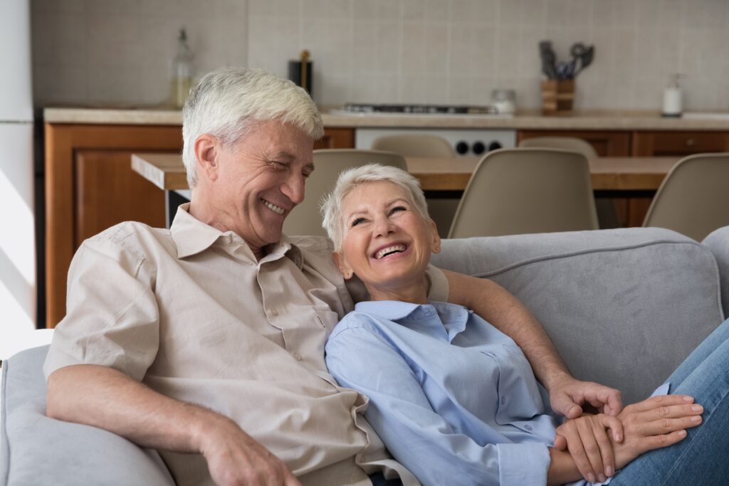 How much do dentures cost? A senior man and woman sit on couch, laughing and looking towards each other happily.