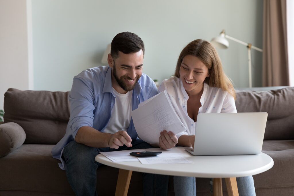 How to choose health insurance in your 30s. A white man and woman sit on couch at coffee table with laptop, bills and calculator. They're smiling.