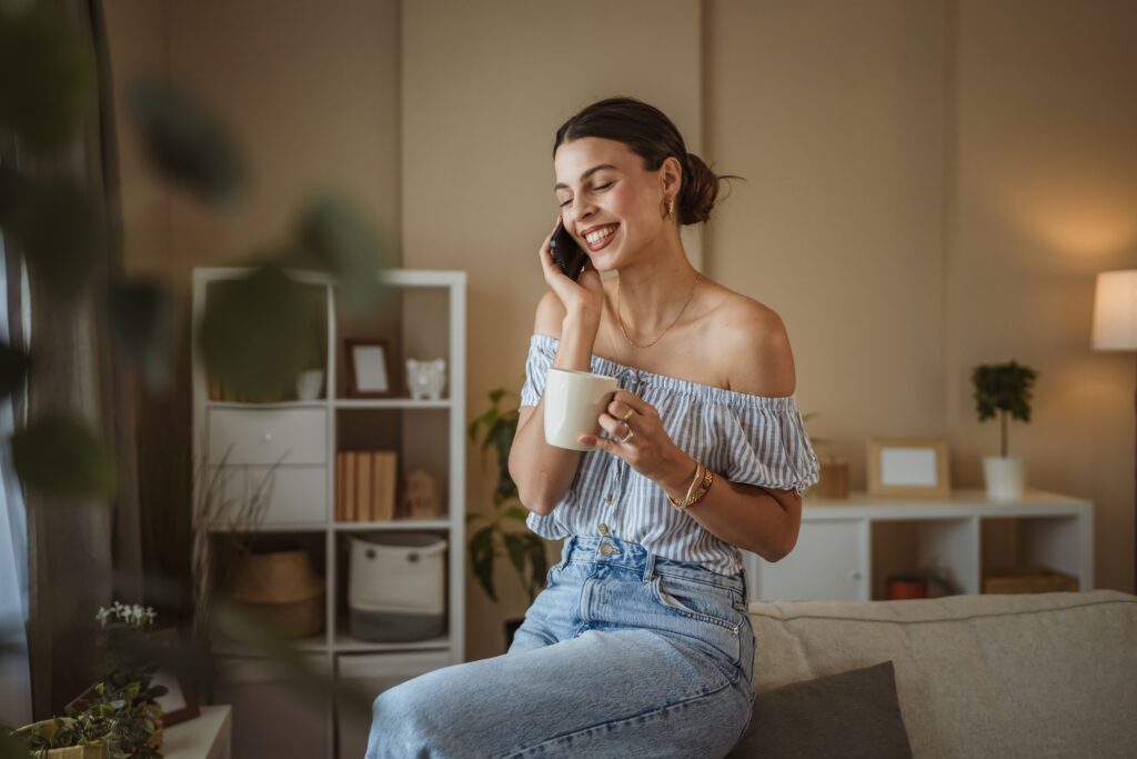 Do I need health insurance in my 30s? A young woman sits on her sofa with a coffee mug, talking on the phone and laughing