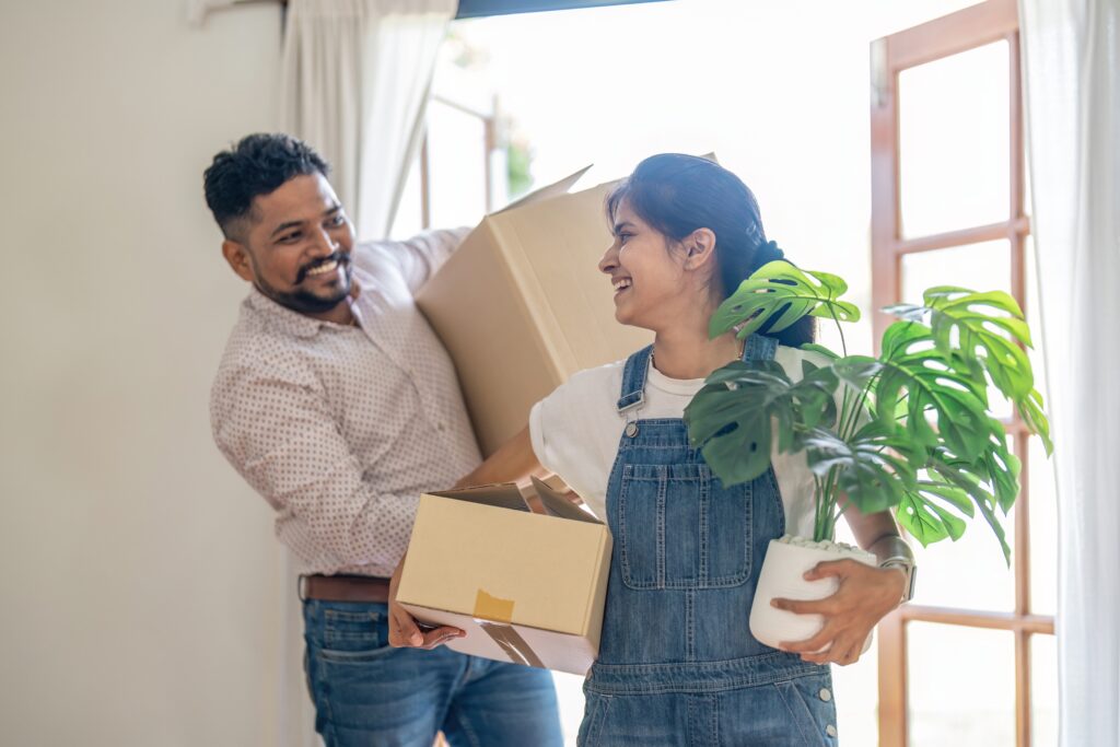 Is couples health insurance cheaper? A couple carry boxes and a plant into their new home.