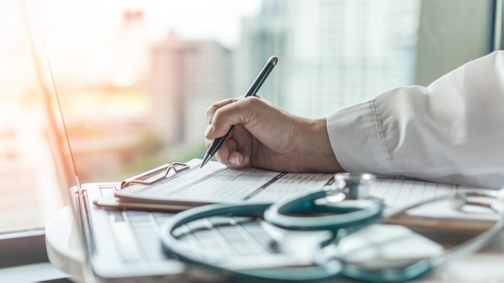 What's the difference between an excess and a co-payment? A male clinician fills out a form with a stethoscope and laptop also on the desk.