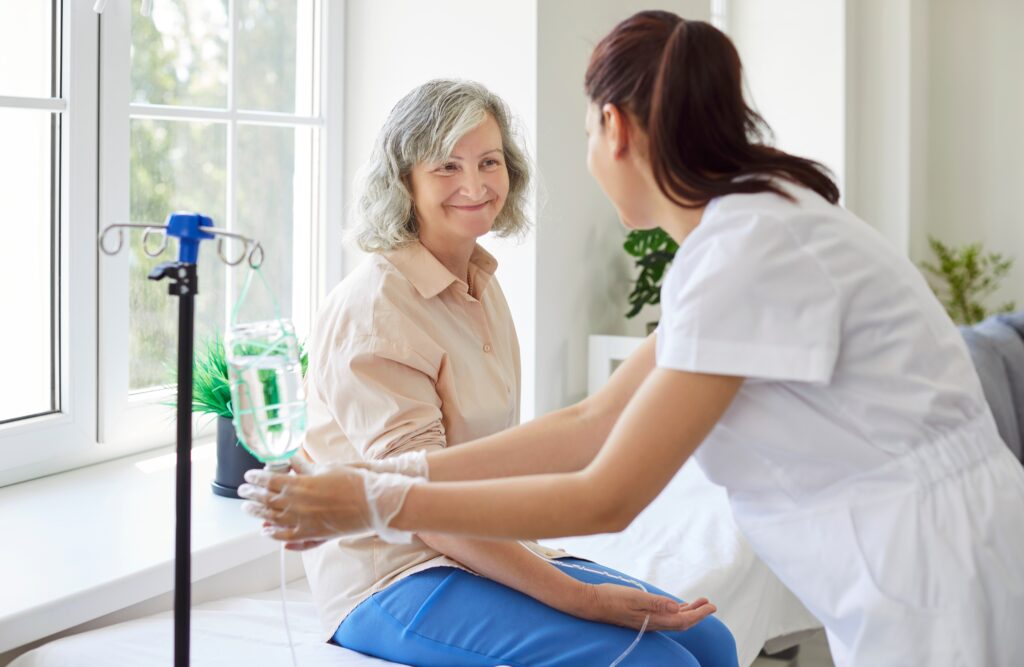 Claiming chemo at home on health insurance. A middle-aged woman smiles at a nurse adjusting her IV in a home setting.