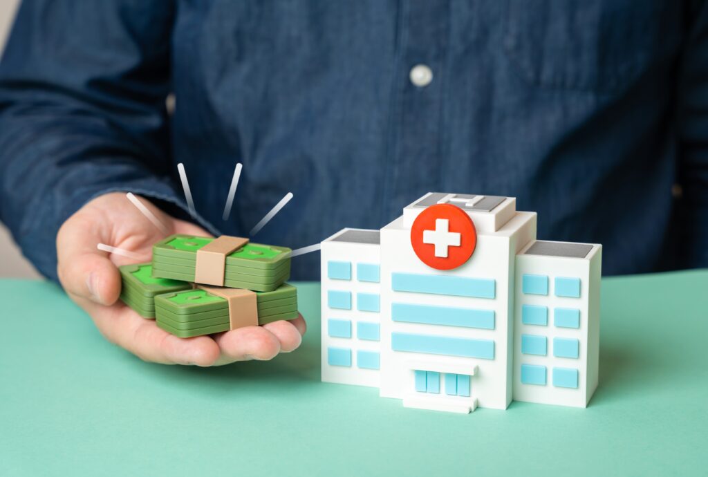 Hospital Cover excess. A man holds piles of small toy money in front of a small toy hospital. 